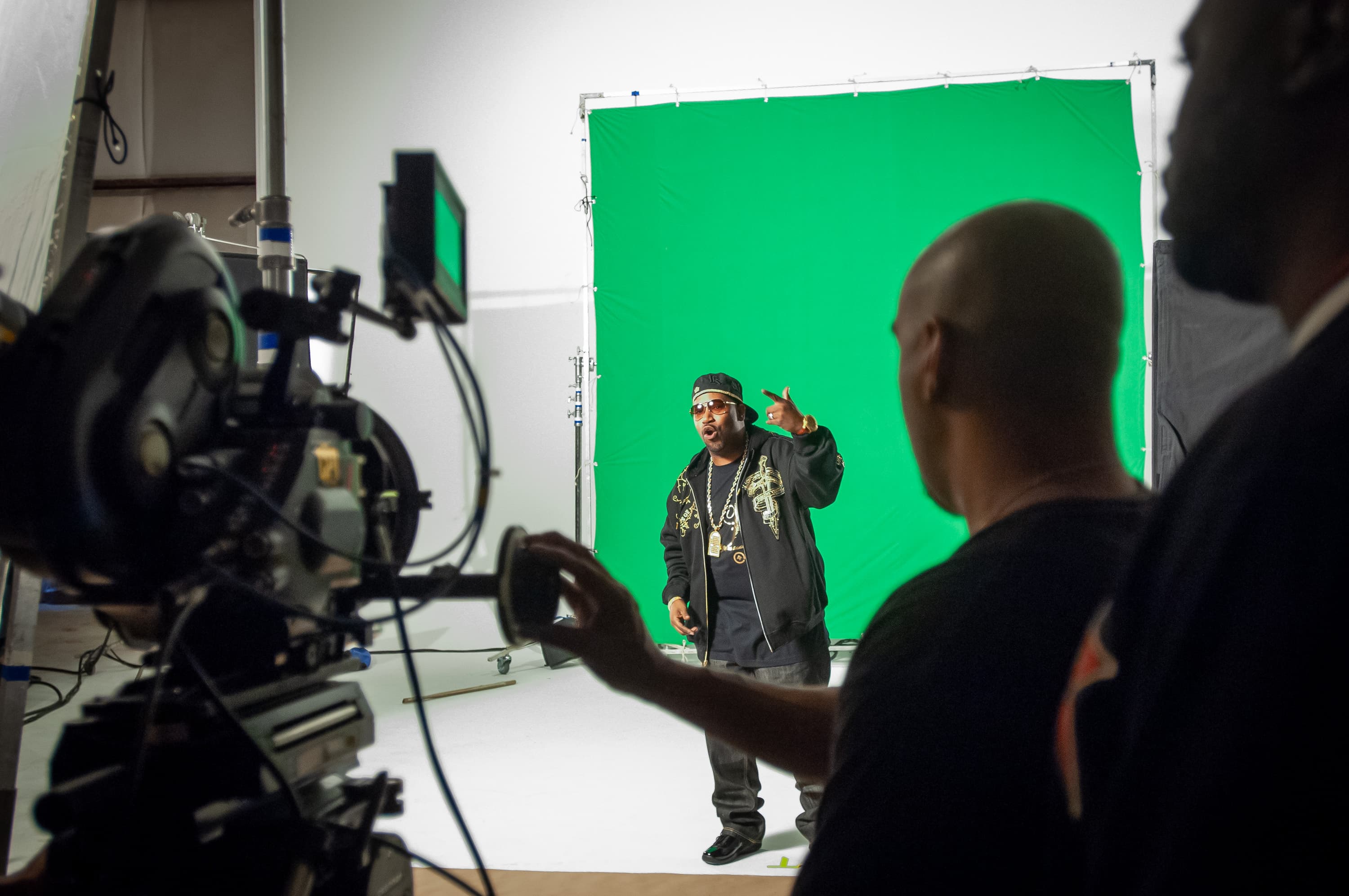 Rapper Bun B standing in front of a green screen while filming a music video, with the camera and assistant camera operator out of focus in the foreground.