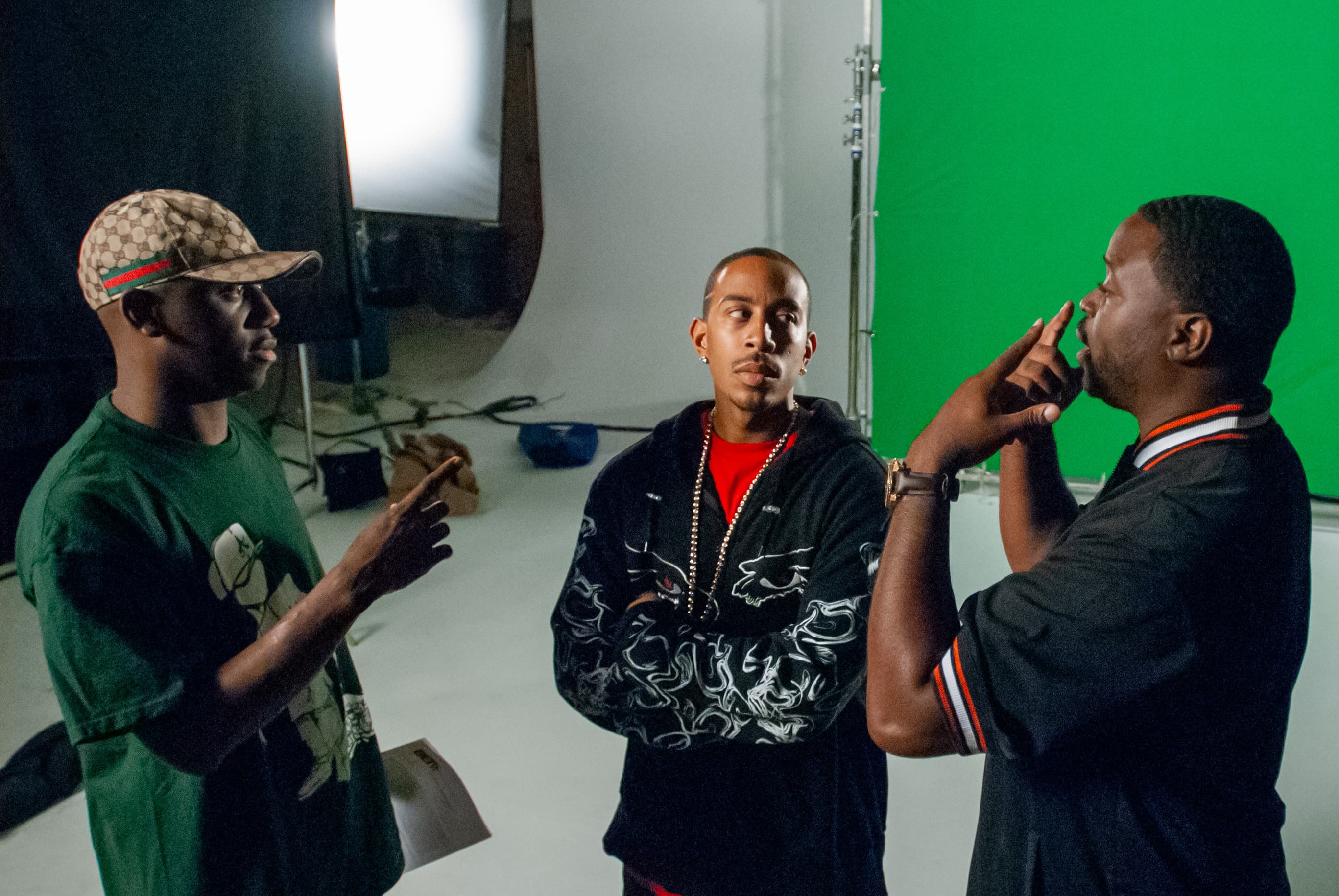 Director Dr. Teeth points to his eyes to demonstrate a move to Ludacris and his manager Chaka Zulu on the set of the  "Down In Tha Dirty" music video. There is a white wall with a screen screen to the right.