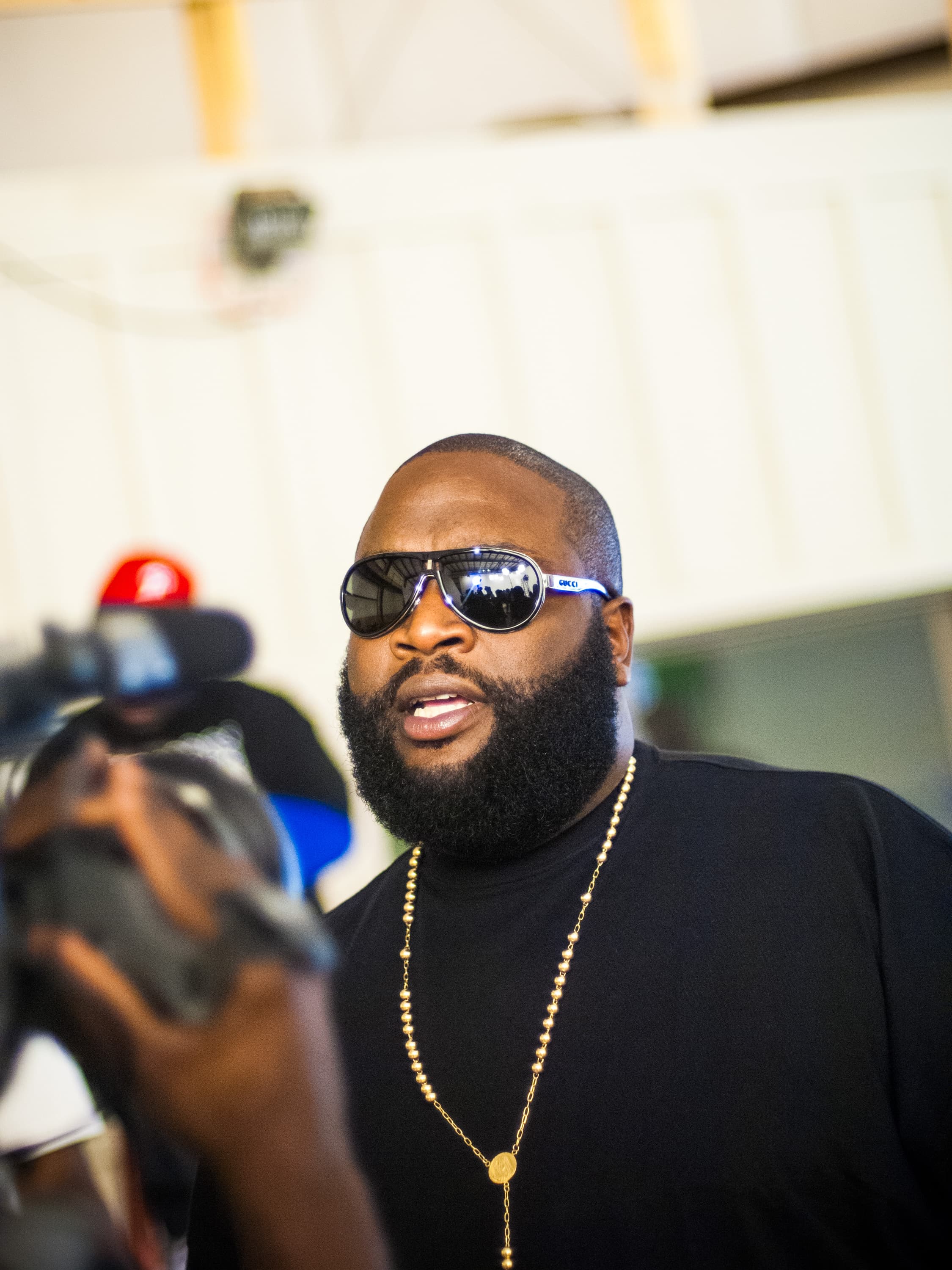 Rapper Rick Ross wearing black shirt, gold chain and dark mirrored sunglasses, speaking to a video camera partially out of frame.