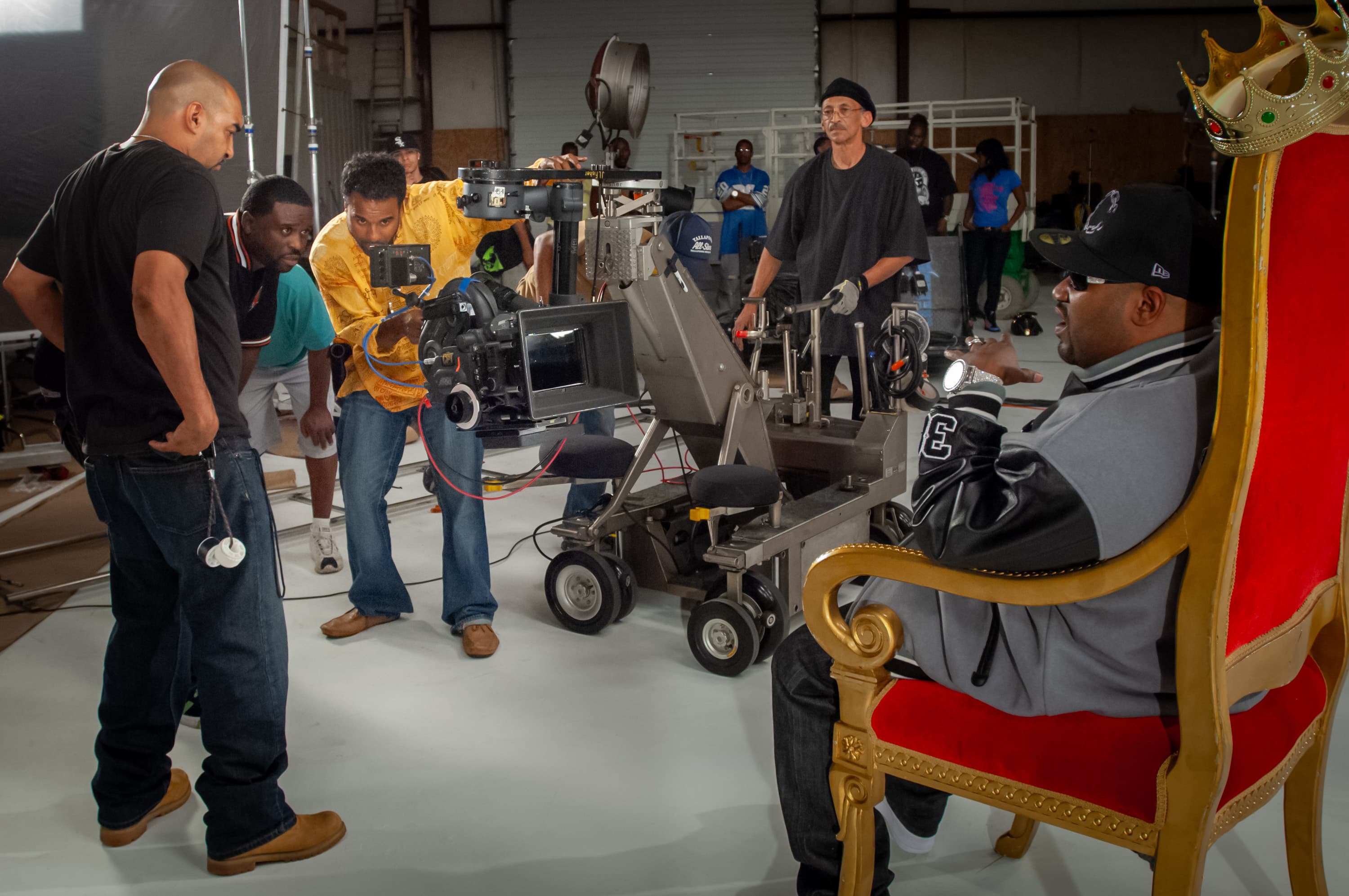 Film crew gathered around a movie camera on a dolly looking at a monitor. The camera is pointed at rapper Bun B sitting in a red velvet chair with gold trim with a plastic crown perched on top.