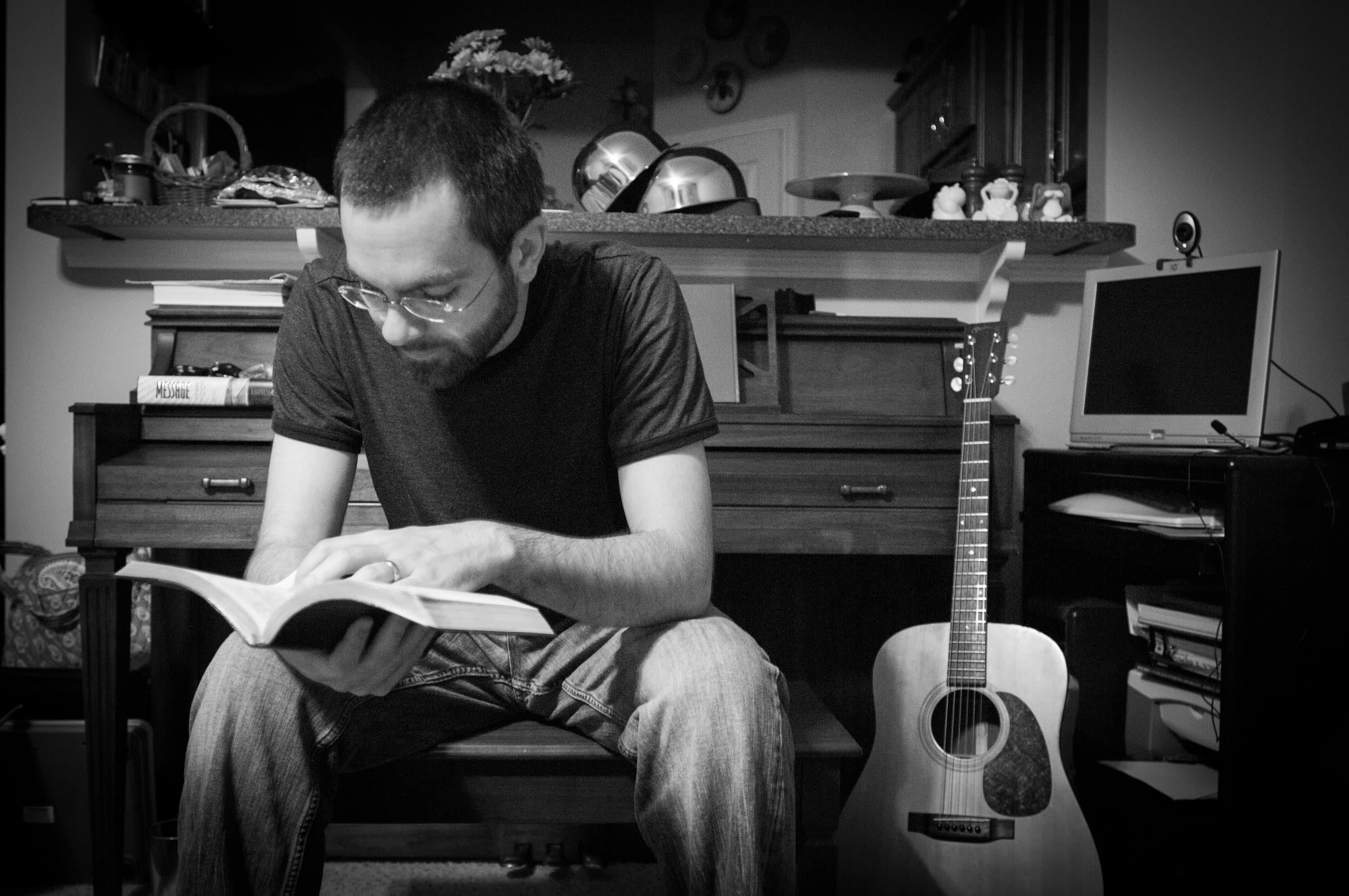 A man sitting on a bench reading a book in a room. A guitar is leaning against the bench, and there is a small computer monitor on a nearby table. Various items are on the countertop behind him, including pots and decorative objects.