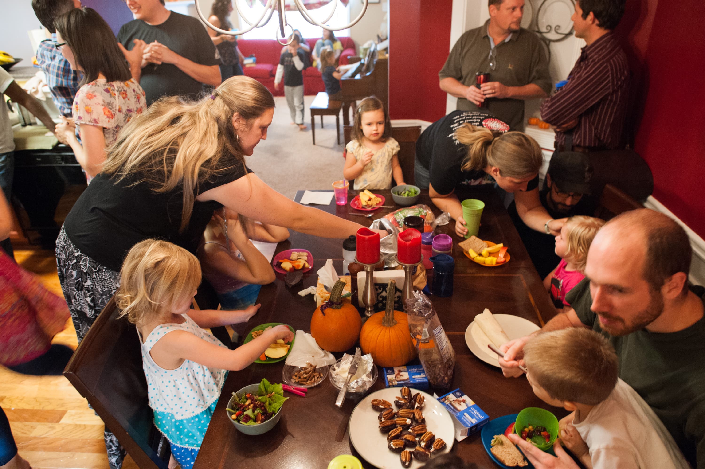 A group of adults and children gathered around a dining table with various foods, including fruits, vegetables, and snacks. Two pumpkins and candles are on the table. People are talking and interacting, and there are more people in the background near a red couch.