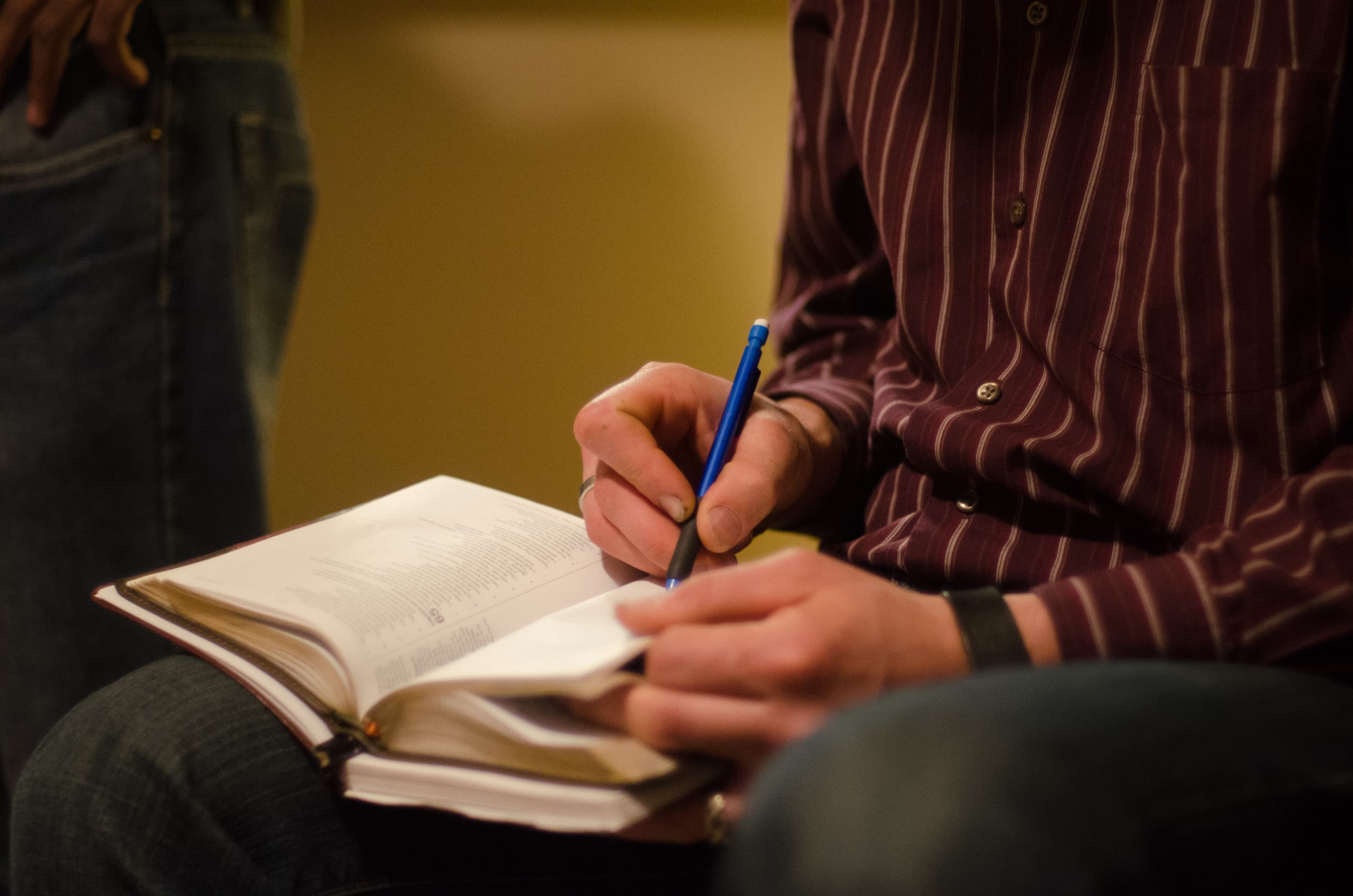 A person wearing a striped shirt is writing with a blue pen on a notebook placed over an open book.