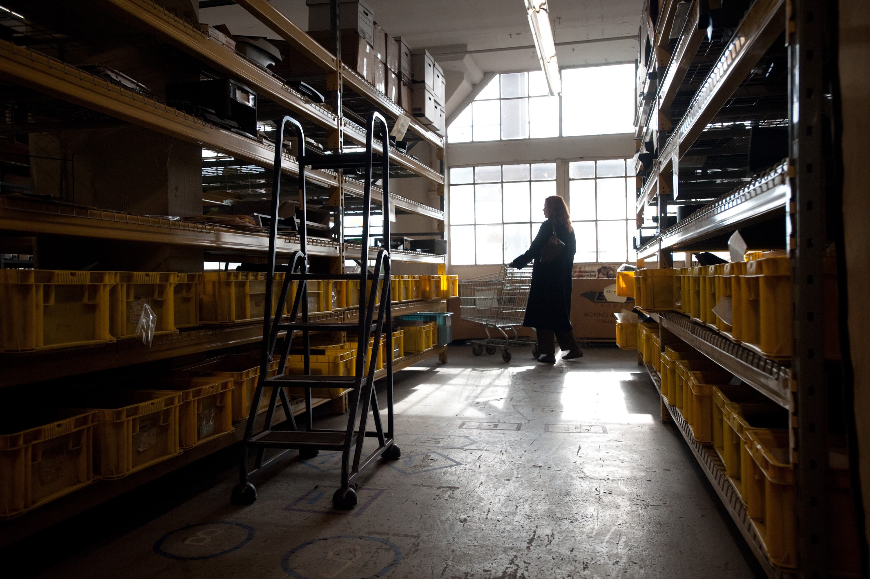 A warehouse with yellow bins on shelves, a woman pushing a shopping cart, and a rolling ladder in the center of the aisle.