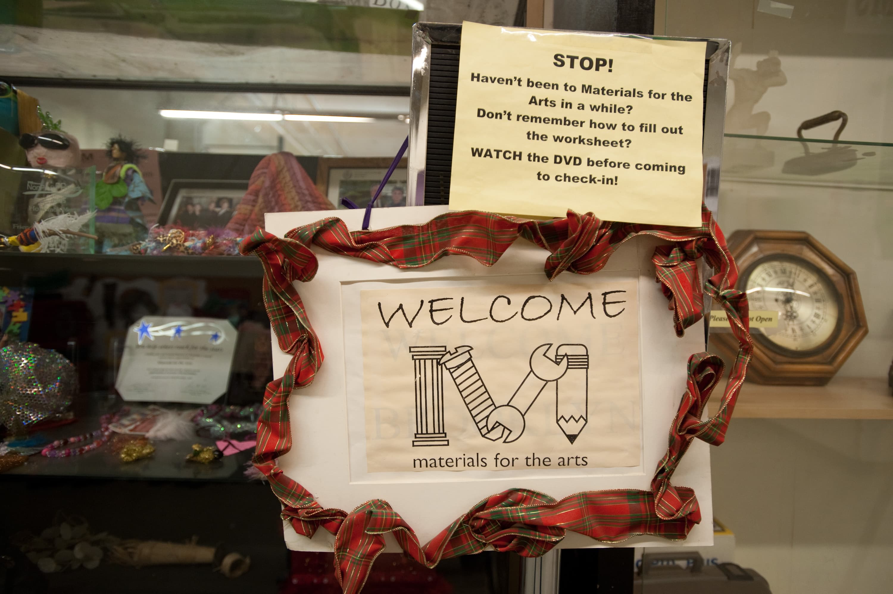 A display featuring a sign with a red ribbon border that reads "WELCOME materials for the arts" with a stylized "M" composed of art tools. Above is a yellow sign with text: "STOP! Haven't been to Materials for the Arts in a while? Don't remember how to fill out the worksheet? WATCH the DVD before coming to check-in!" In the background, there are shelves with decorative items and an antique clock.