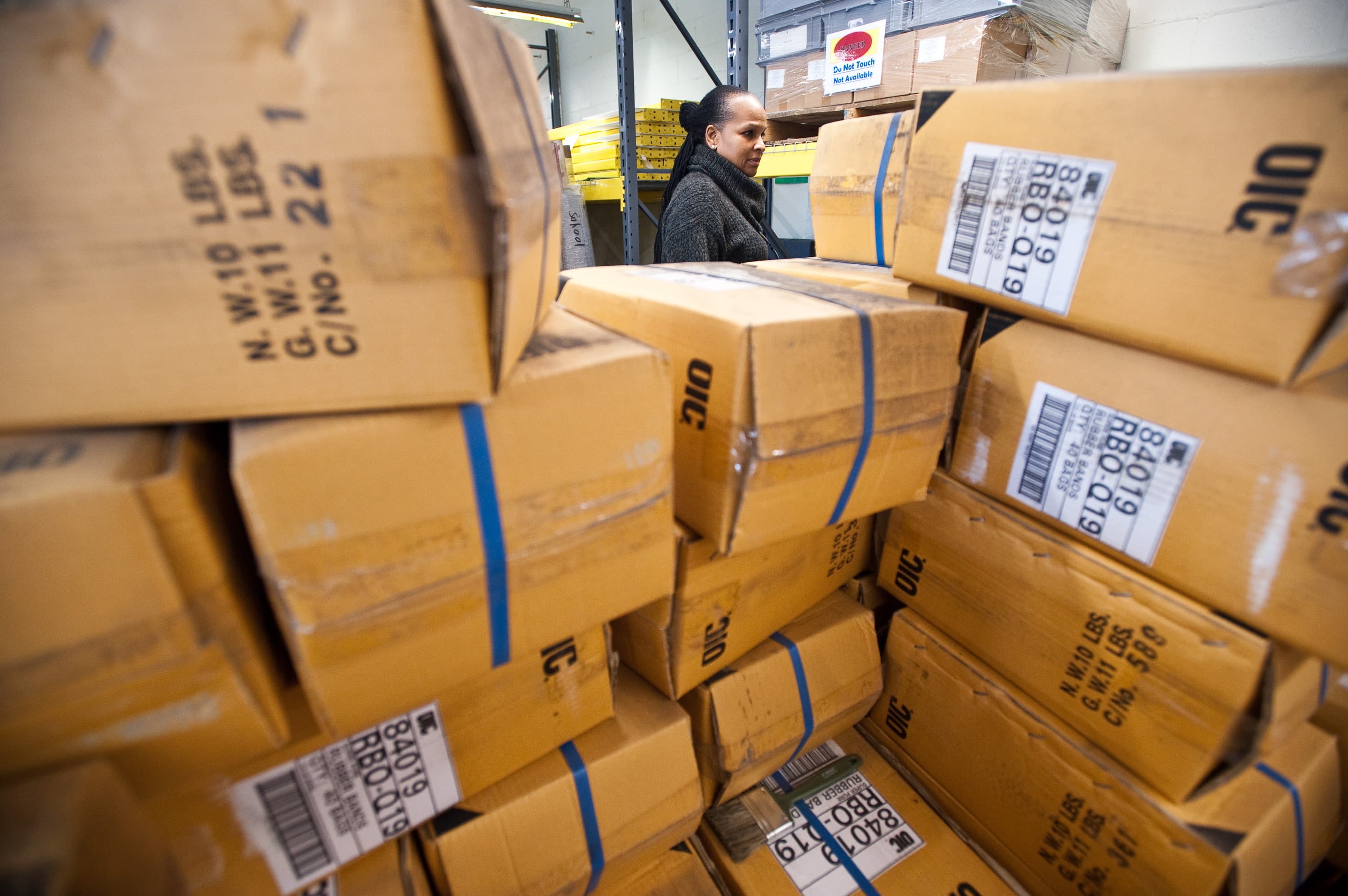 A person standing in a storage area with stacked cardboard boxes. The boxes have labels and are secured with blue straps. A sign in the background reads "Do Not Touch Not Available."