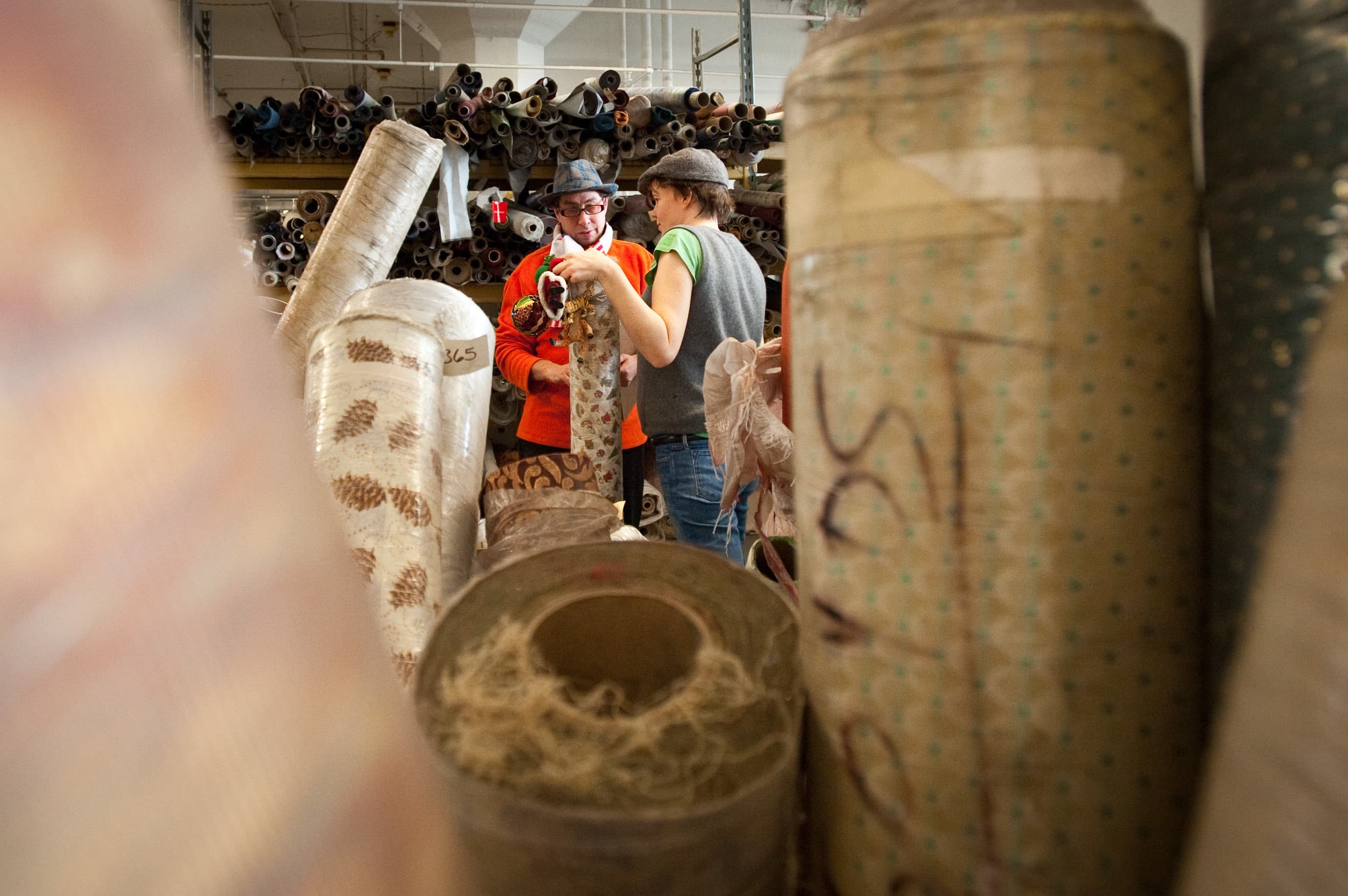 Two people examining a roll of fabric in a warehouse filled with large rolls of textiles.