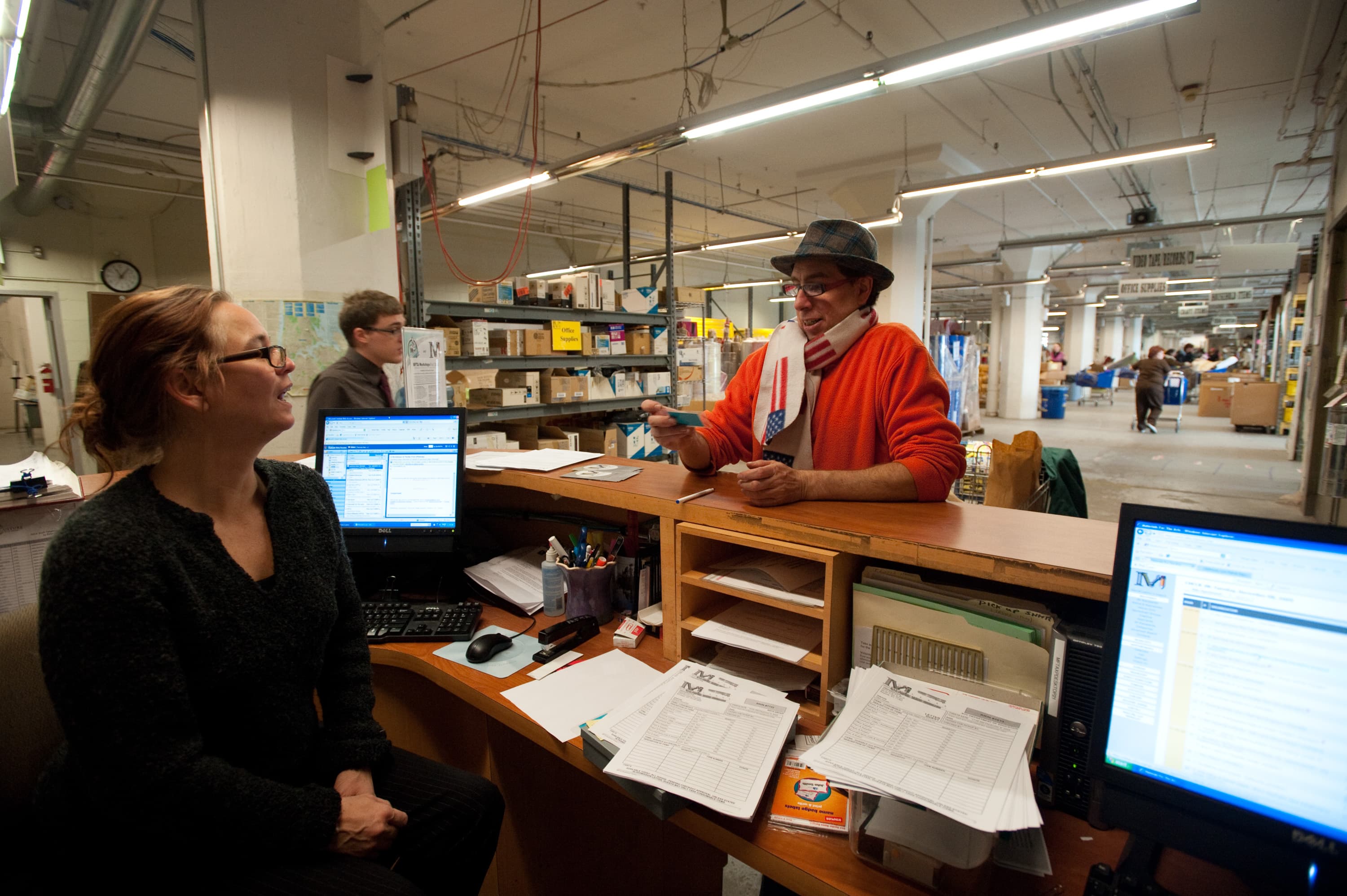 A woman at a desk with two computer monitors in a warehouse or office space interacting with a man in an orange sweater and scarf. Shelves filled with various boxes and items are visible in the background with people working.