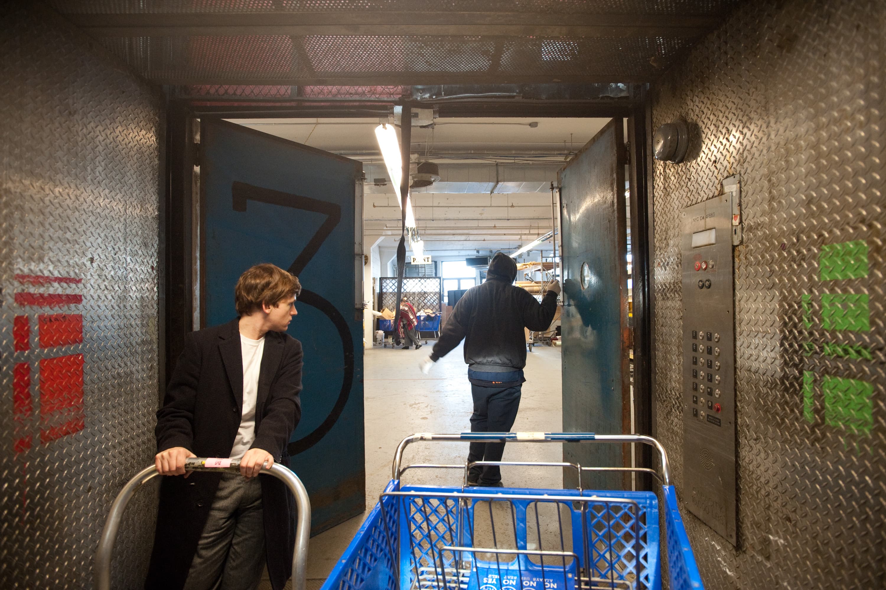 Two people in an industrial setting. One person is pushing a blue shopping cart and looking to the left. Another person is walking through a large blue door marked with the number 3, wearing dark clothing and a hood. The area has textured metal walls and an elevator control panel.