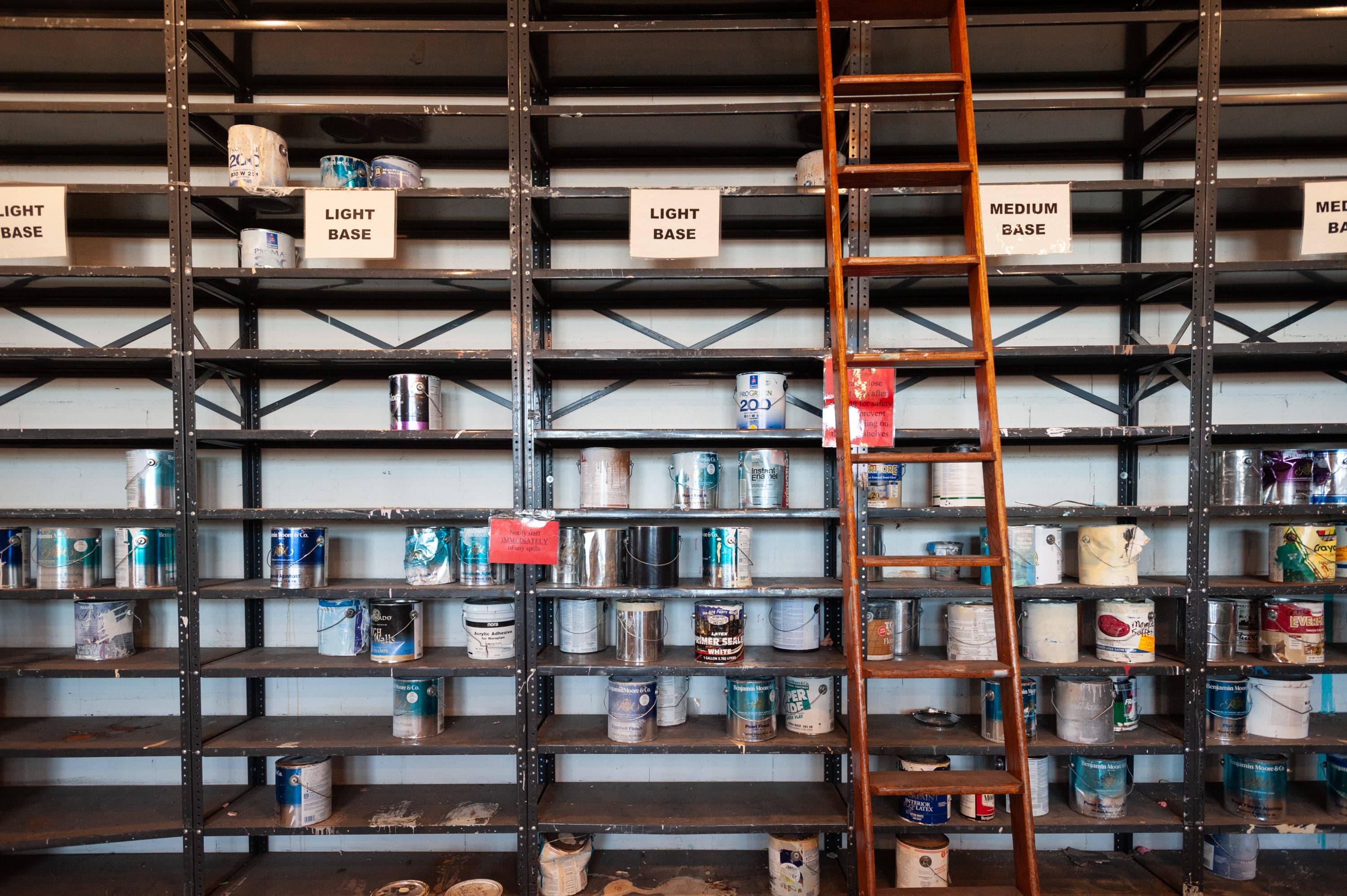 Shelves filled with various paint cans organized by base type. Three sections are labeled "LIGHT BASE" and "MEDIUM BASE". A wooden ladder is leaning against the shelves.