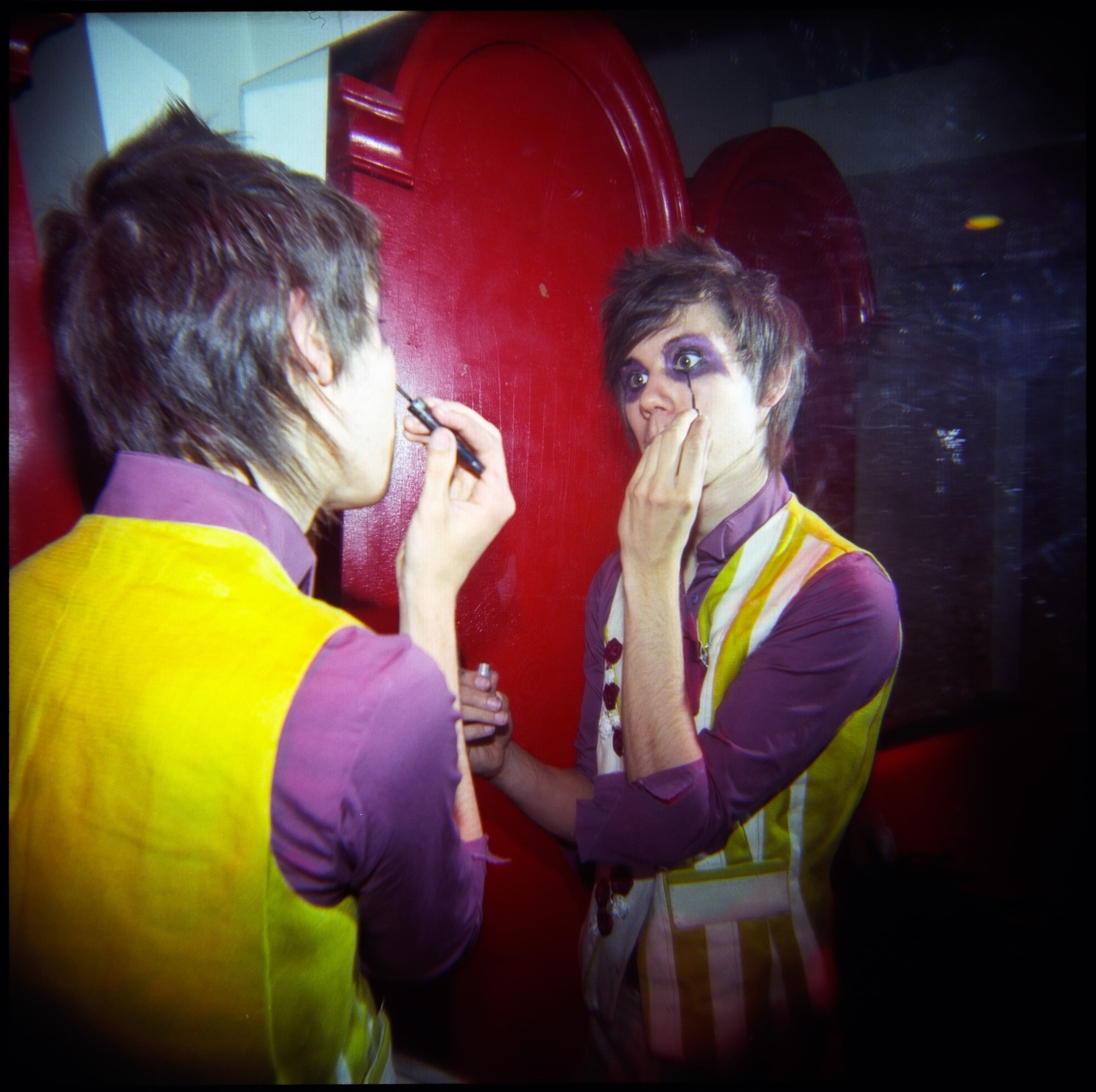 A person applying eye makeup in front of a mirror. They are wearing a yellow and purple outfit and are holding a makeup brush and a small container. The mirror reflects their image and the red background.