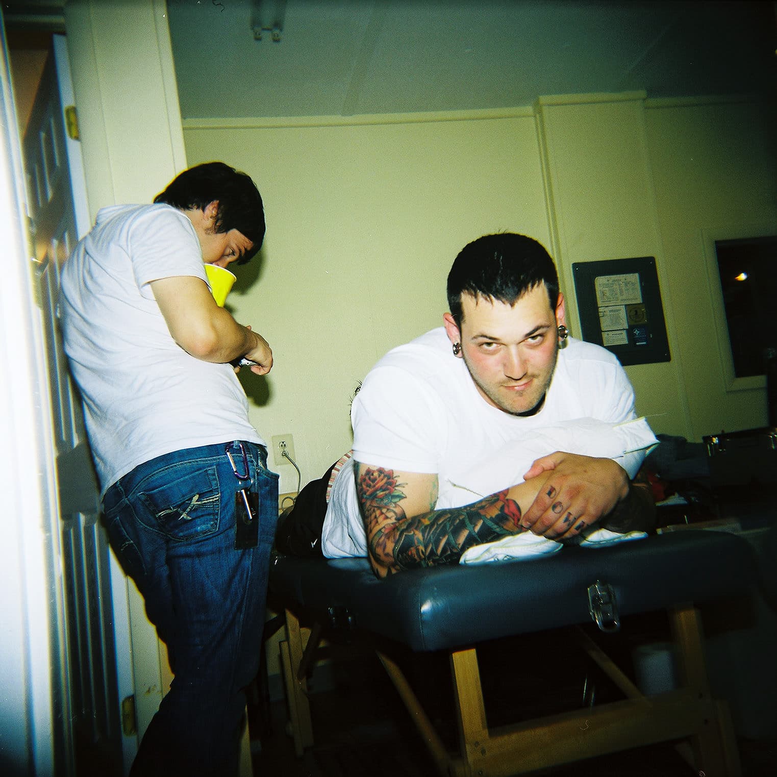 A person with tattoos lies on a table, another person beside them holds a yellow object, possibly engaged in tattooing or another activity in a room.