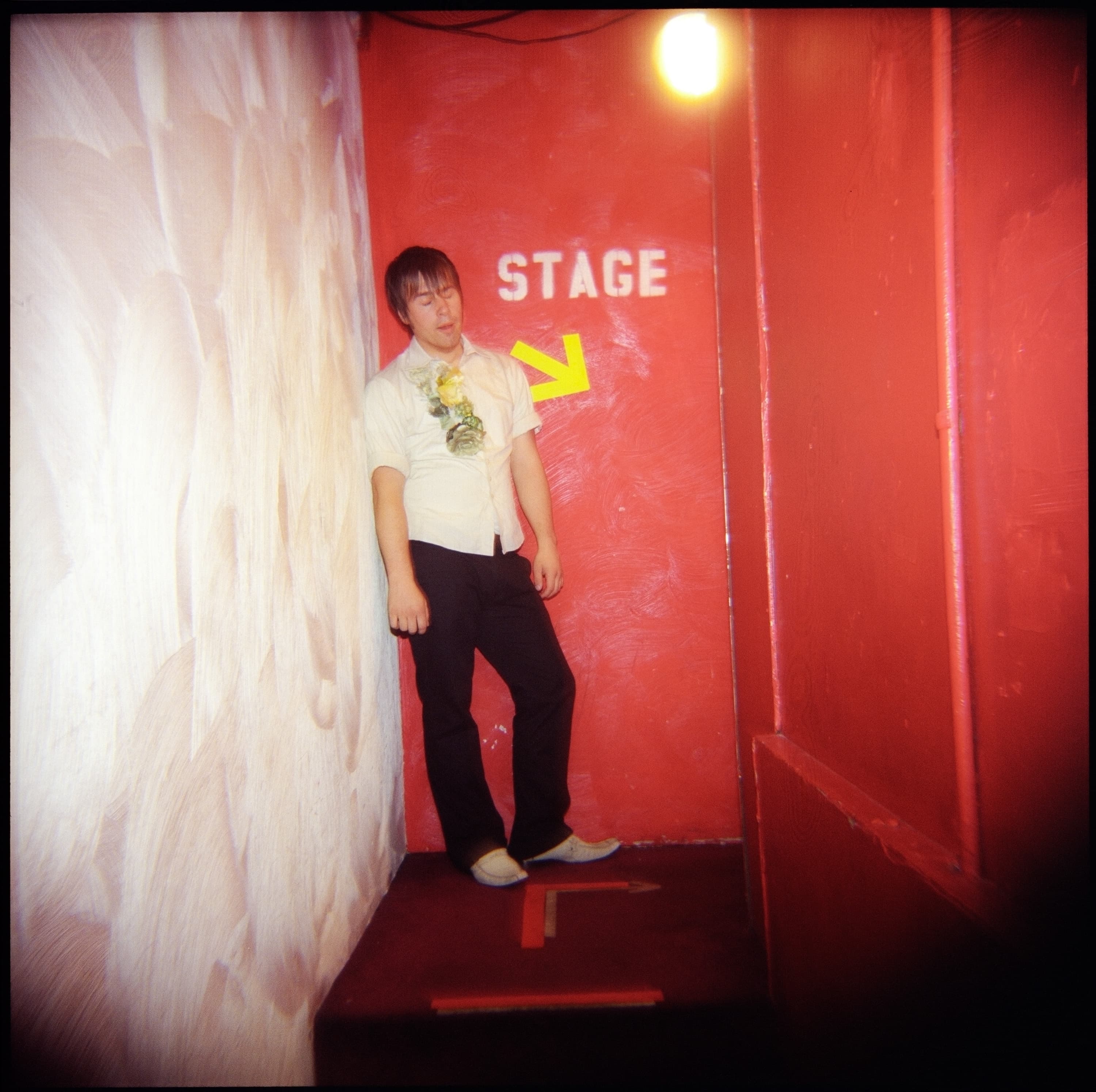 A person leaning against a wall in a narrow hallway. The wall is painted red, and there is a yellow arrow pointing right below the word "STAGE." The person is wearing a light-colored shirt with flowers on the front and dark pants.