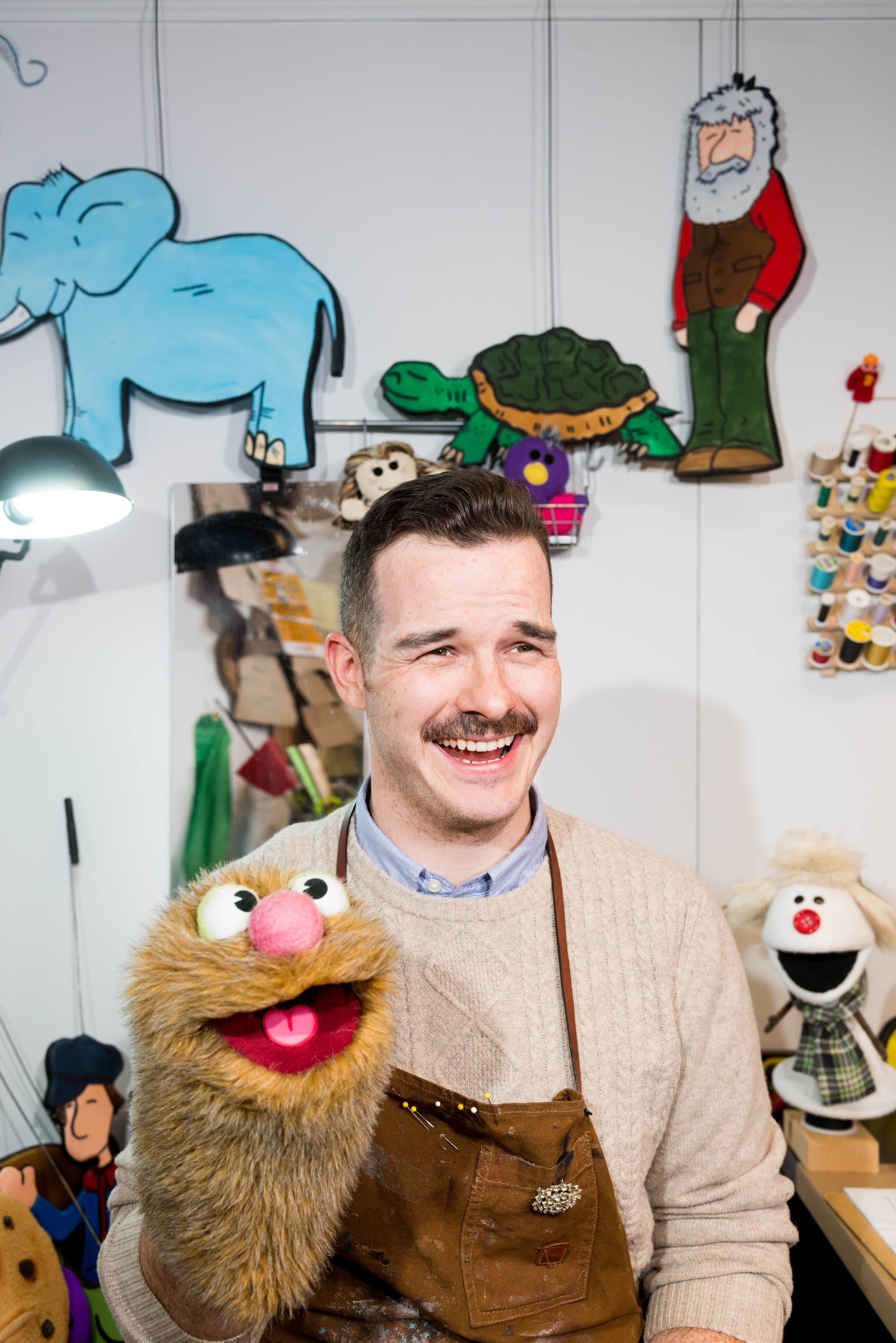 A person holding a puppet with a mustache, wearing an apron. Behind them, colorful cartoon drawings of an elephant, a turtle, and a bearded figure are visible on the wall. Spools of thread and other crafting materials are also seen in the background.