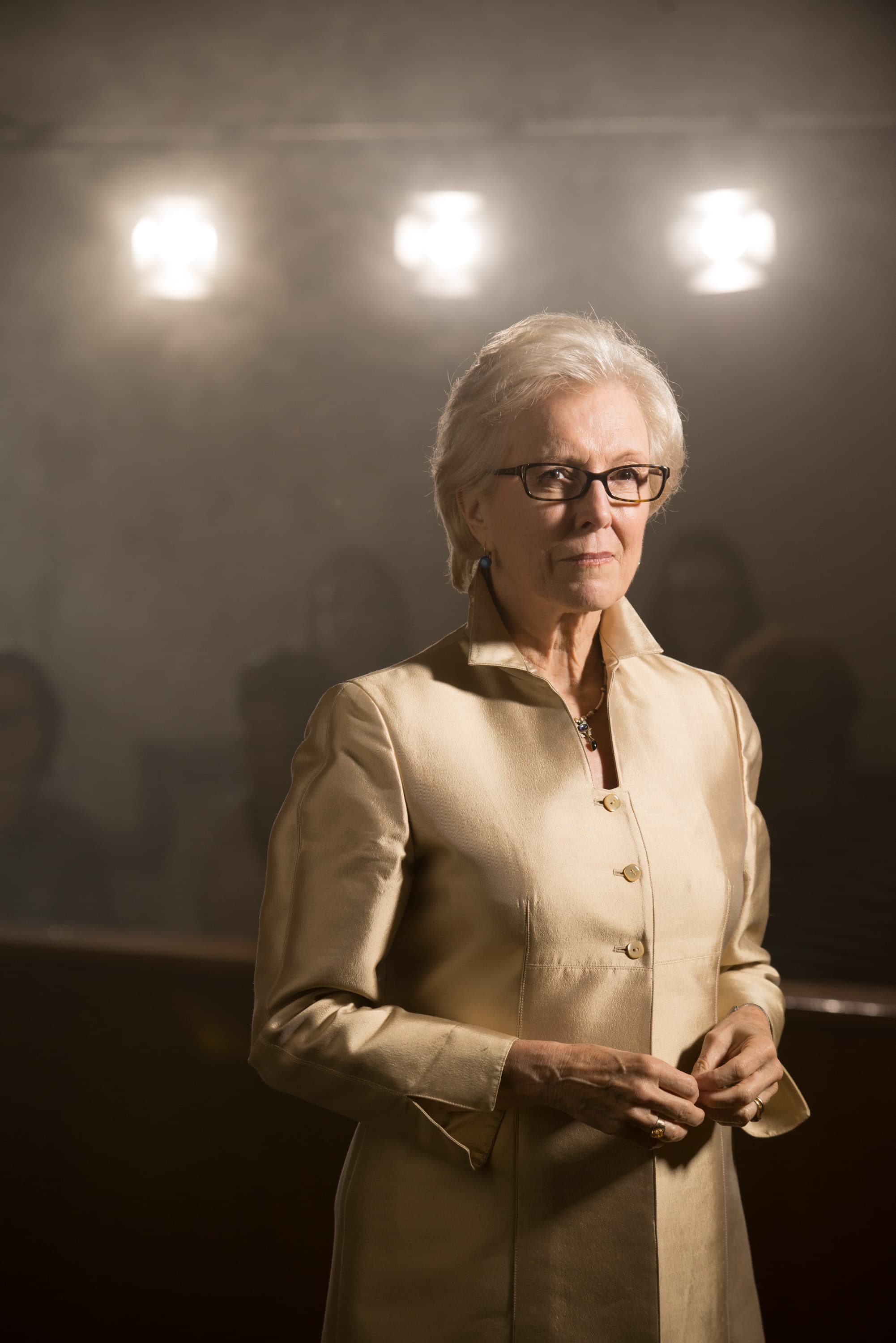 An older woman wearing glasses and a light-colored jacket stands with her hands clasped. She is in a softly lit indoor setting with three lights in the background.