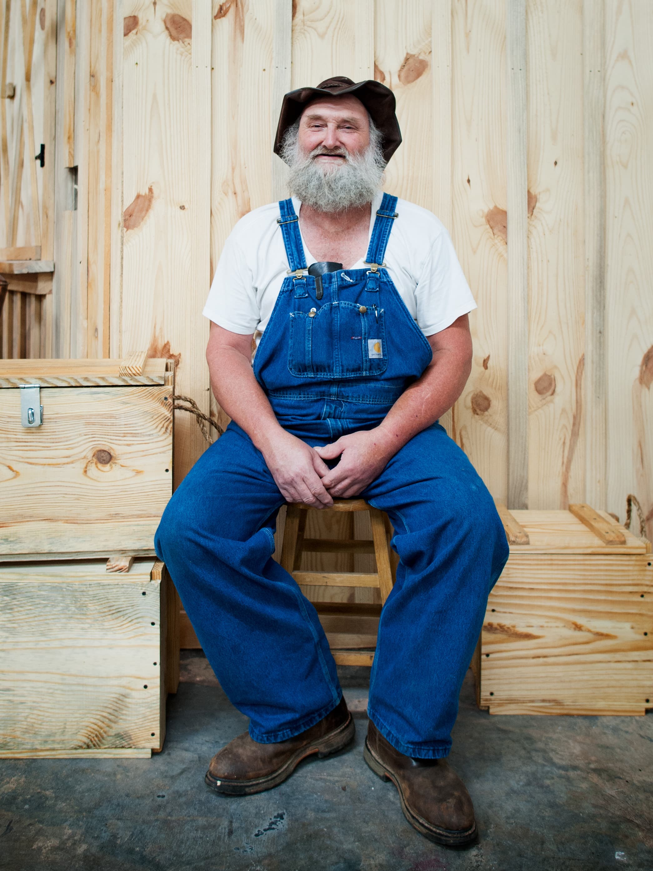 A bearded man wearing a brown hat, white t-shirt, and blue denim overalls, sitting on a wooden stool in front of a wooden wall with several wooden crates nearby.