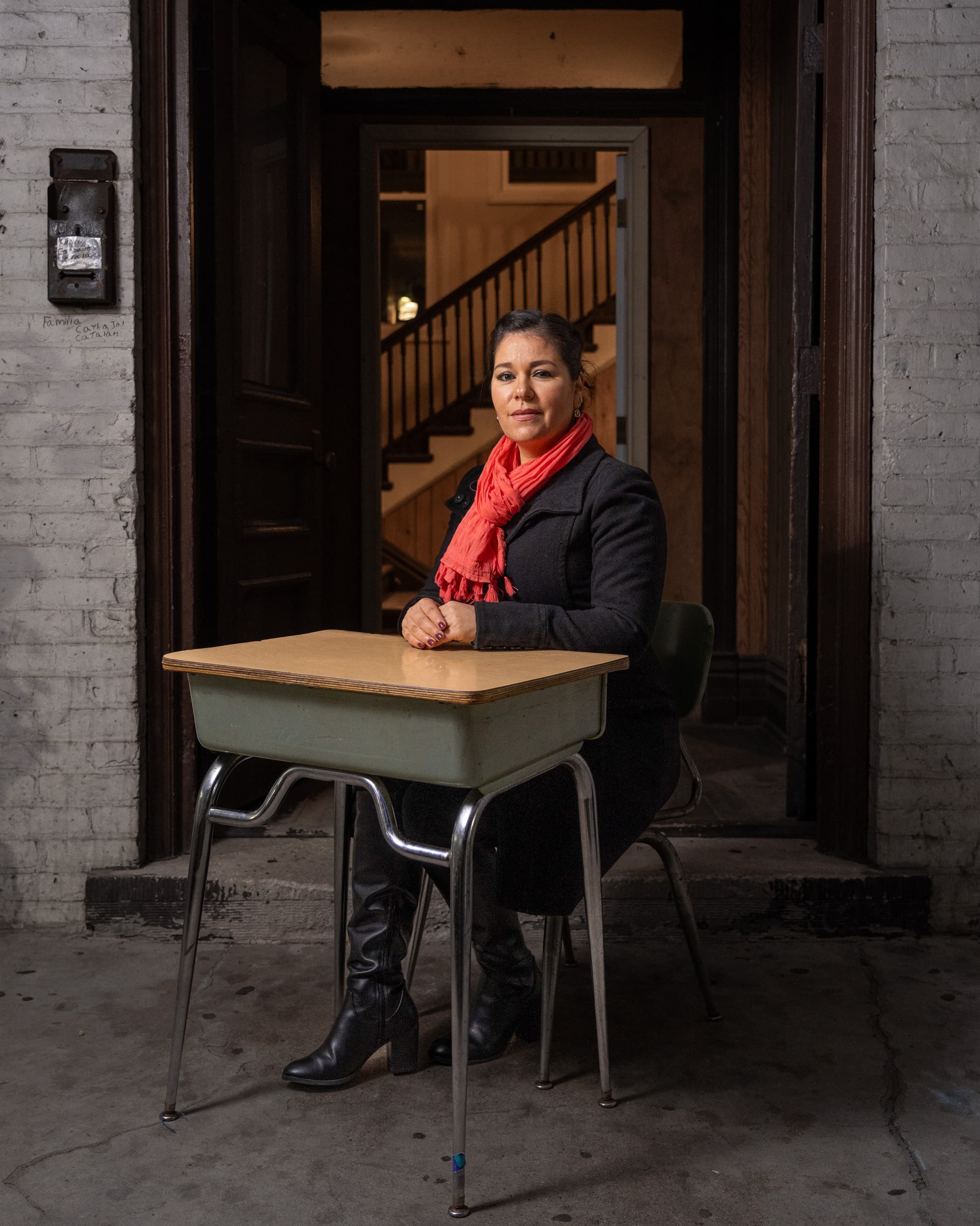 A person sitting at a school desk inside a building with visible stairs in the background. They are wearing a red scarf and black boots.