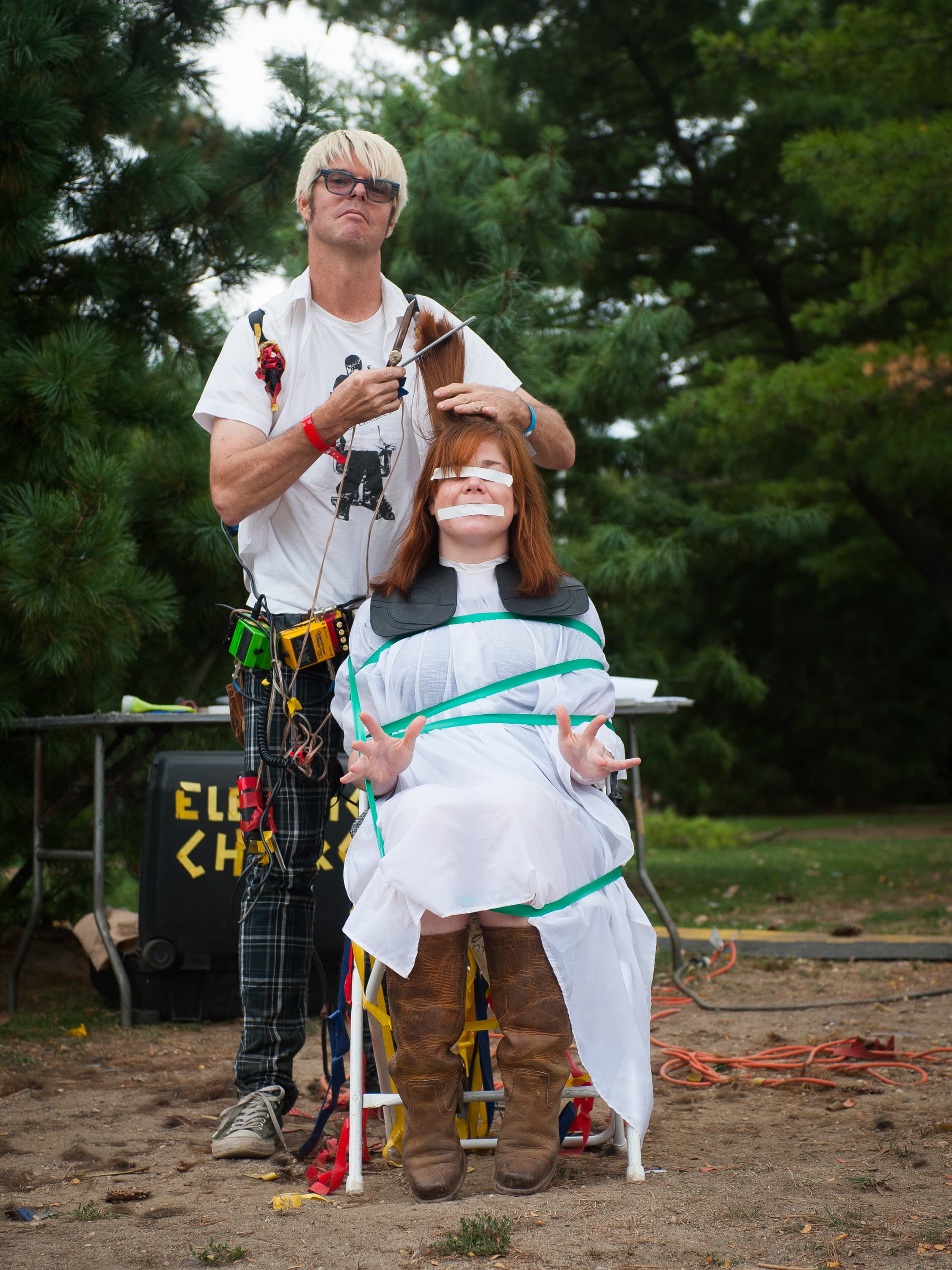 A man with blond hair and glasses is cutting a woman's hair outdoors. The woman is sitting on a chair, wrapped in a sheet and restrained with green tape. Behind them is a table with a sign partially visible, reading "ELE... CH..."