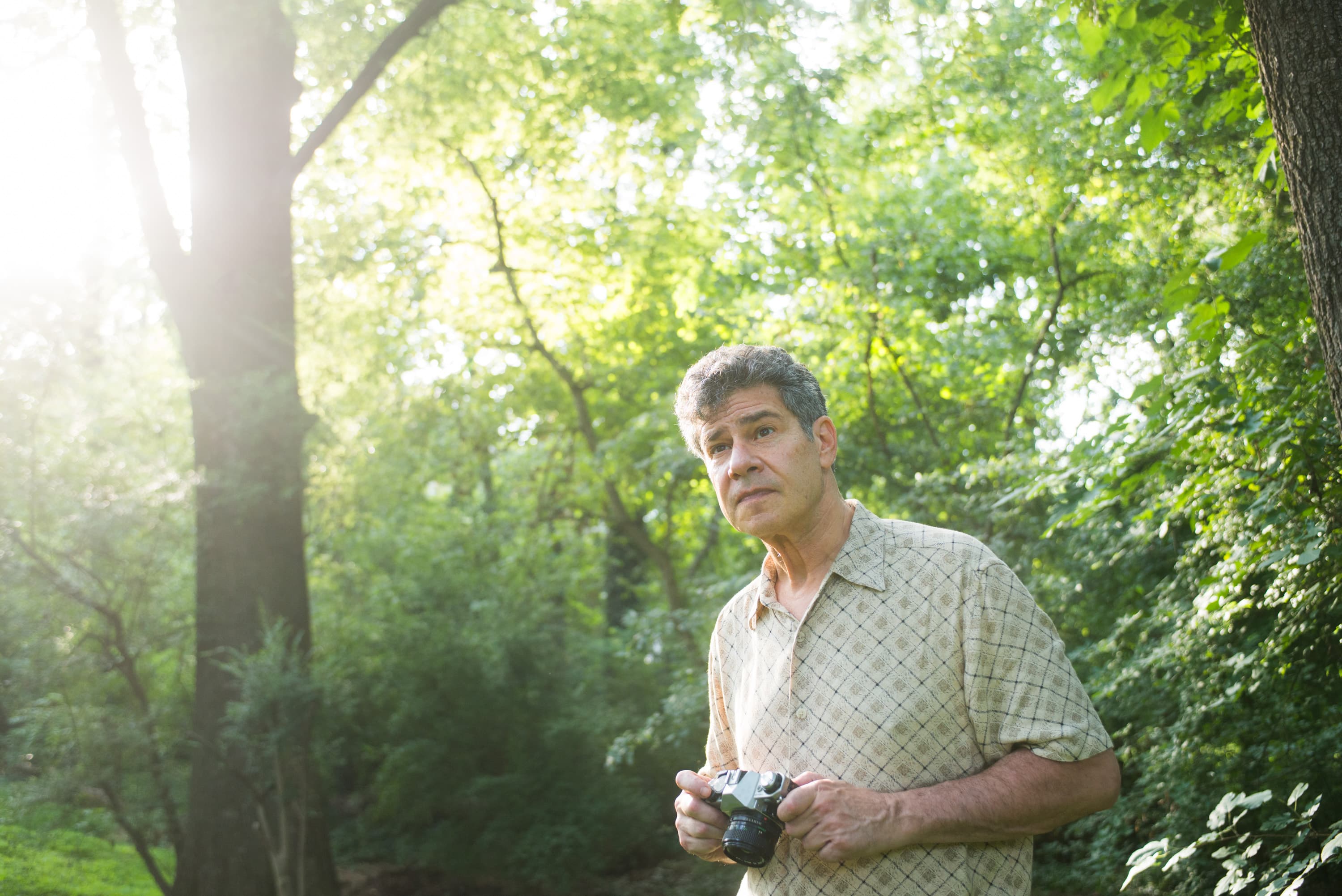 A man holding a camera stands in a forested area, surrounded by trees and greenery.