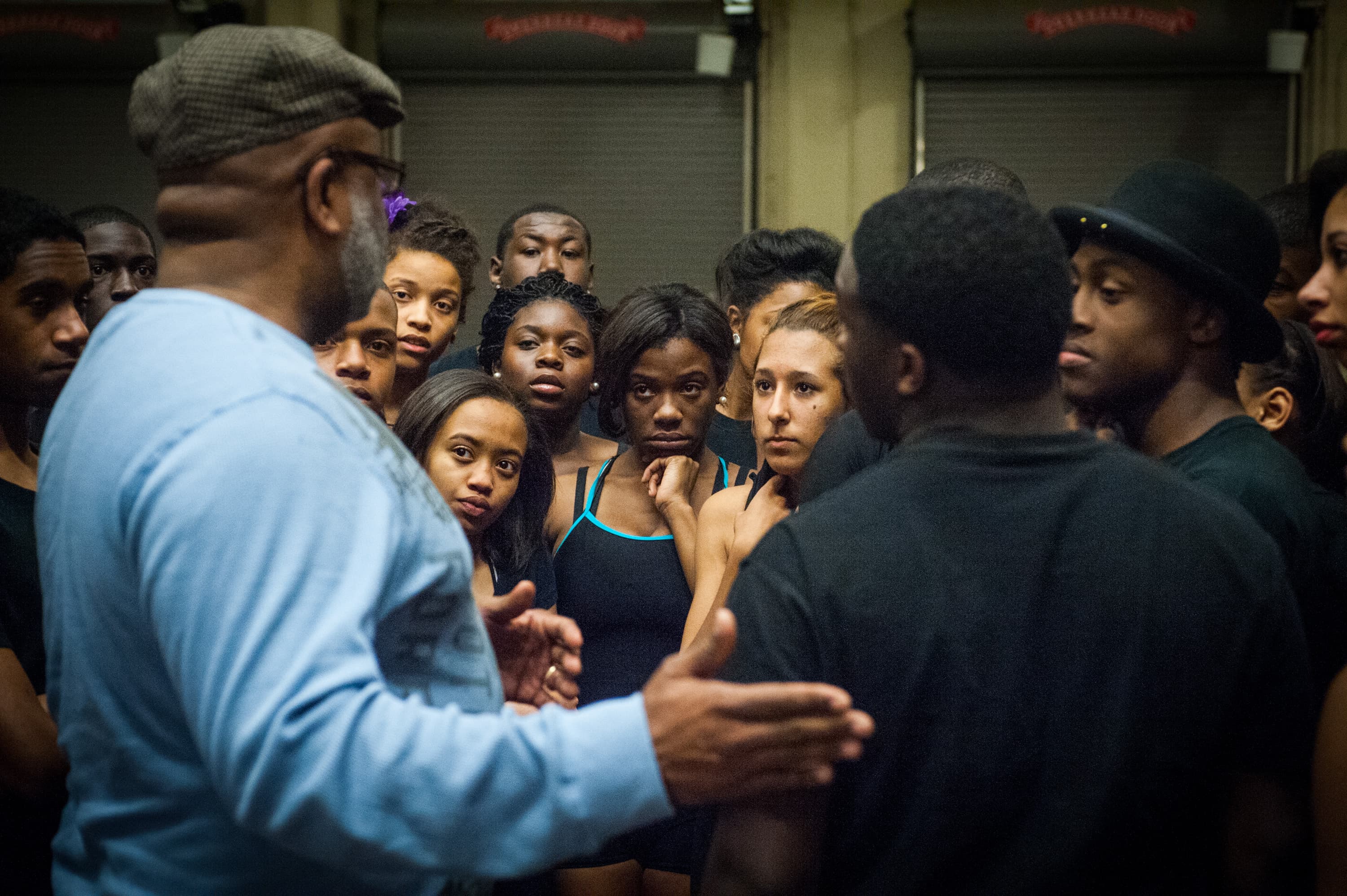 A group of people gathered closely together, with a man in a blue shirt and a hat speaking to them. The group appears attentive and is composed mostly of young people.