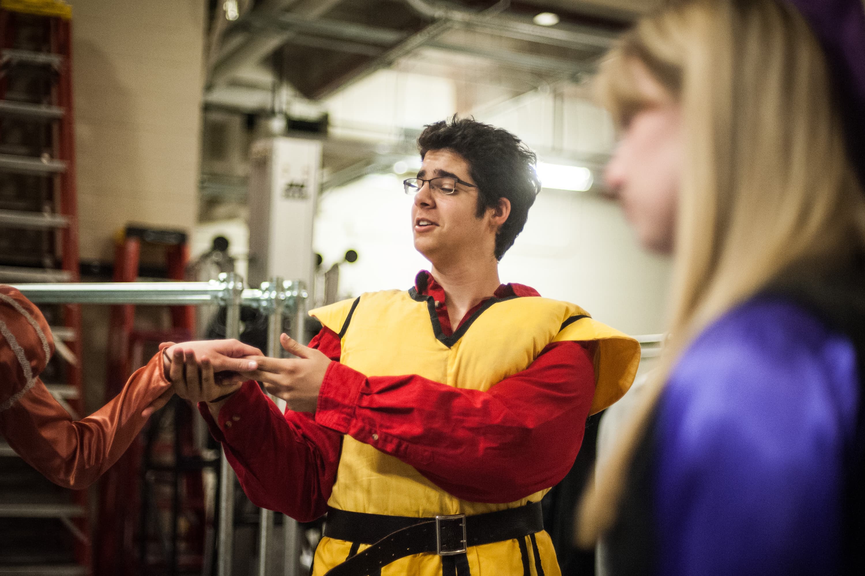 A person wearing a yellow and red costume, holding hands with another person dressed in a similar style. They appear to be in a backstage or industrial setting with ladders and metal structures visible in the background.