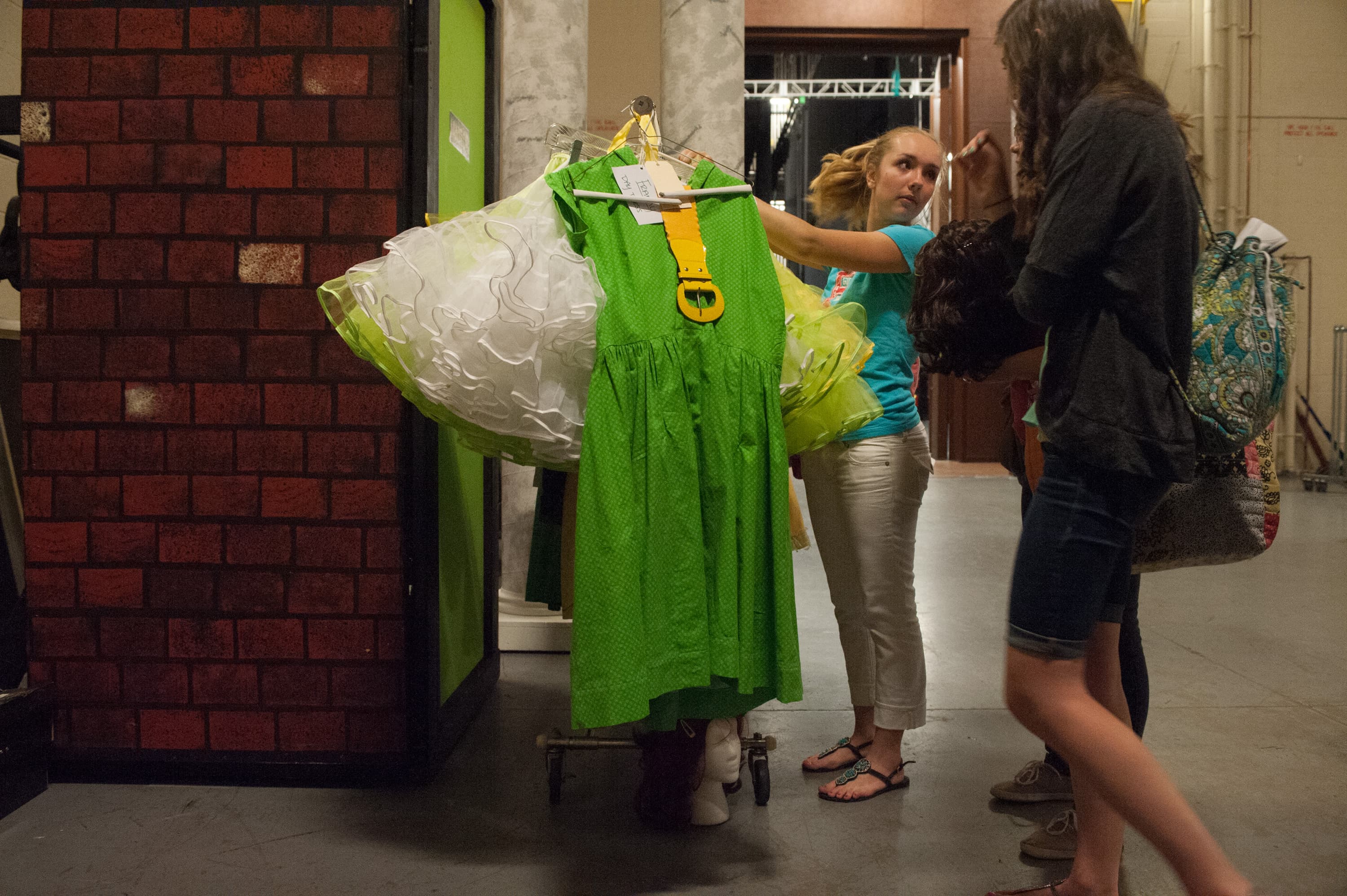 Two people examining a bright green dress on a hanger backstage. Part of a costume rack with other garments is visible, along with a mannequin head on the floor.