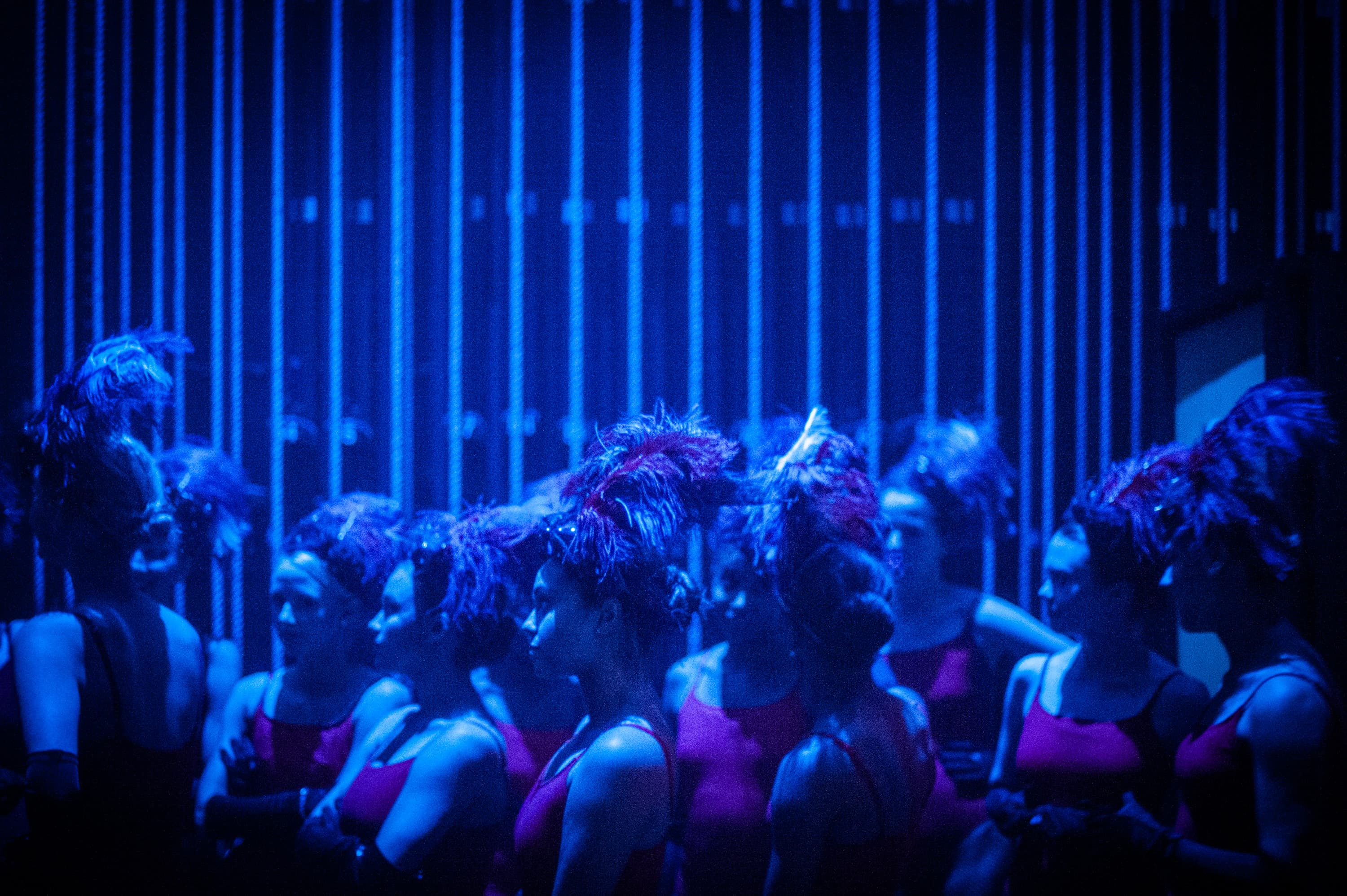 A group of performers in red costumes with feathered headpieces standing backstage in blue lighting.