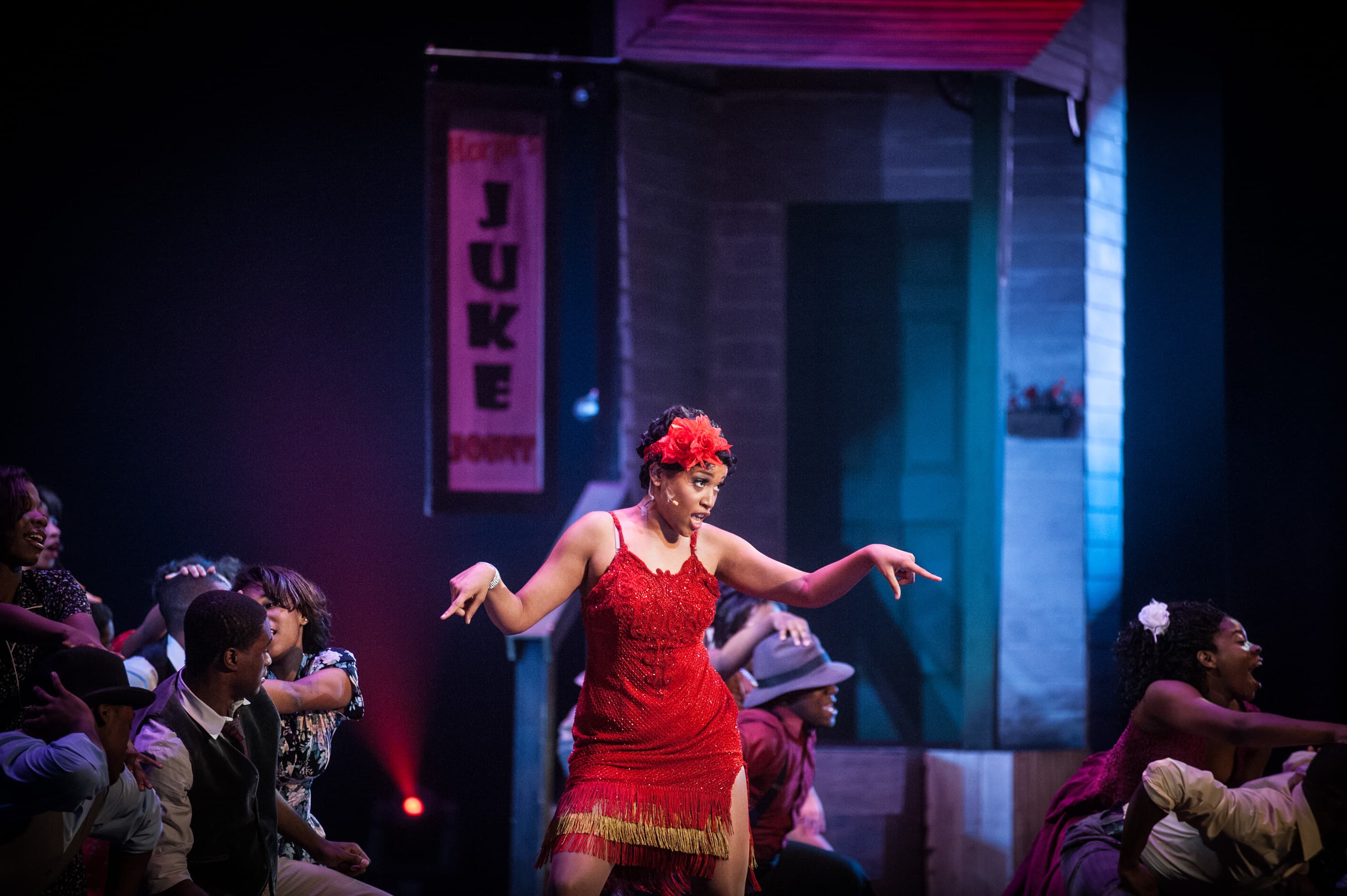 A performer in a red dress dances on stage, surrounded by other dancers. A sign in the background reads "Juke Joint."