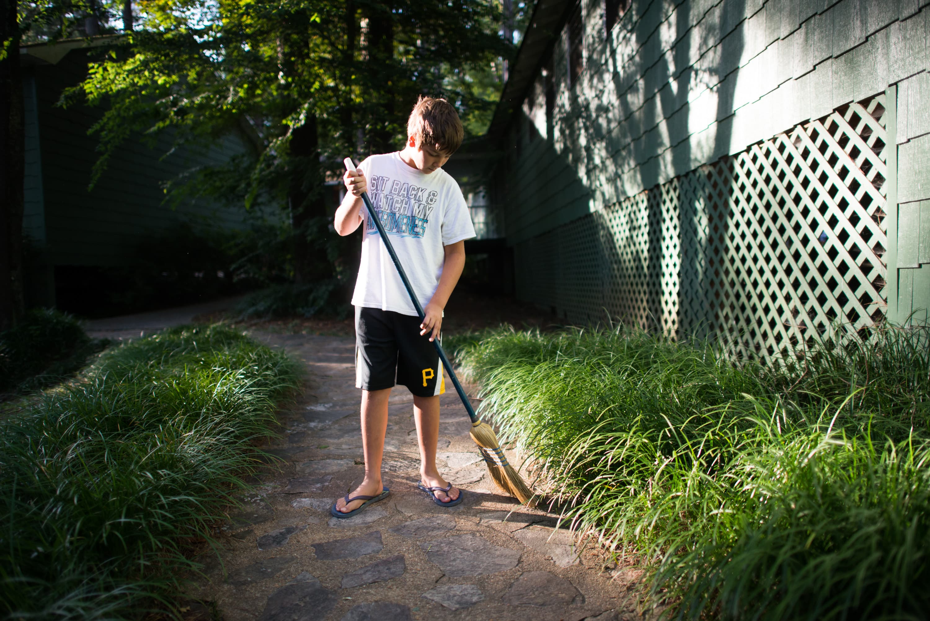 A young person in a white shirt and black shorts is sweeping a stone path surrounded by grass. The person is wearing flip-flops and a building with latticework is visible in the background.