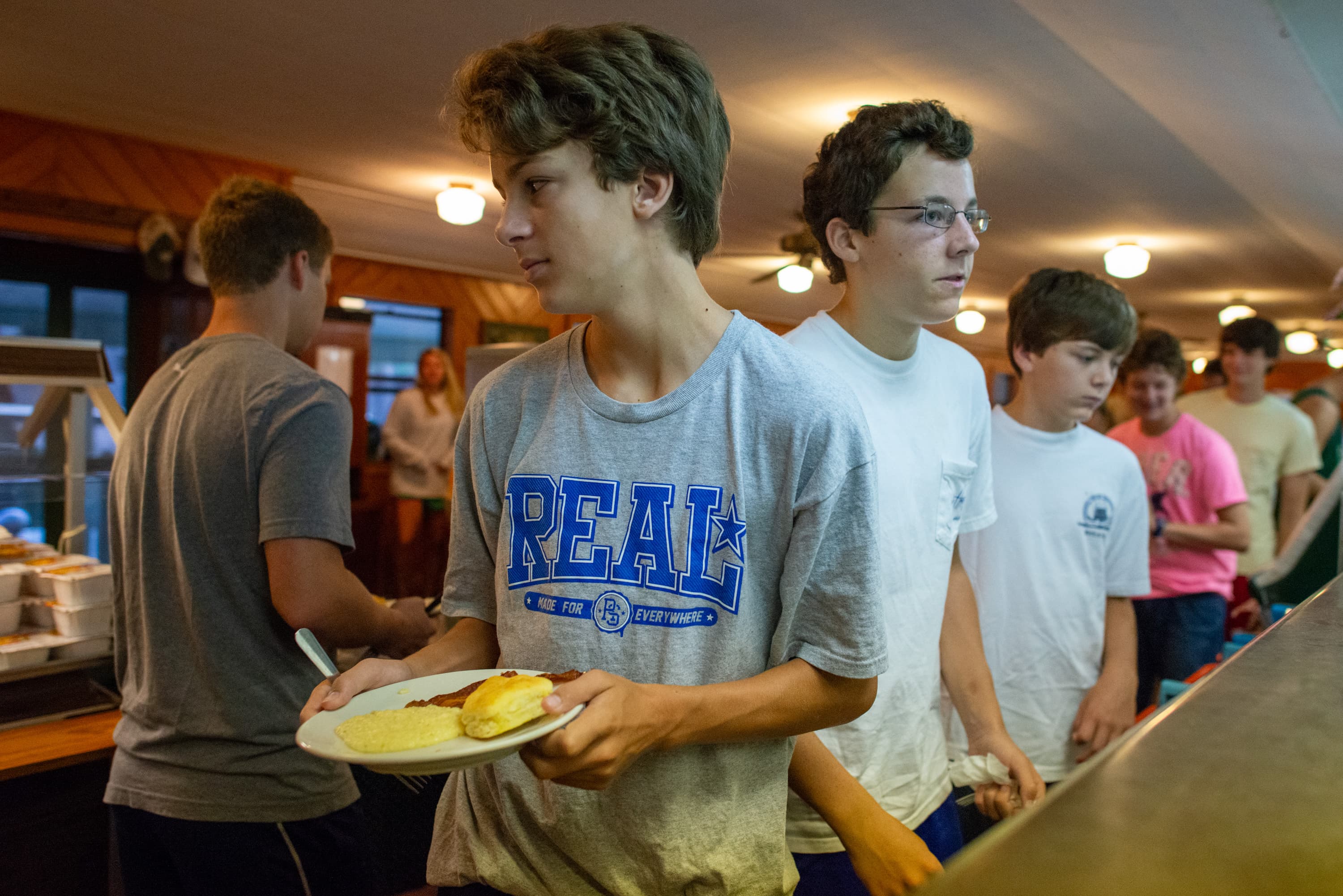 A group of young people standing in line at a cafeteria. One person in the foreground is holding a plate of food. The person is wearing a gray shirt with the word 'REAL' printed on it.