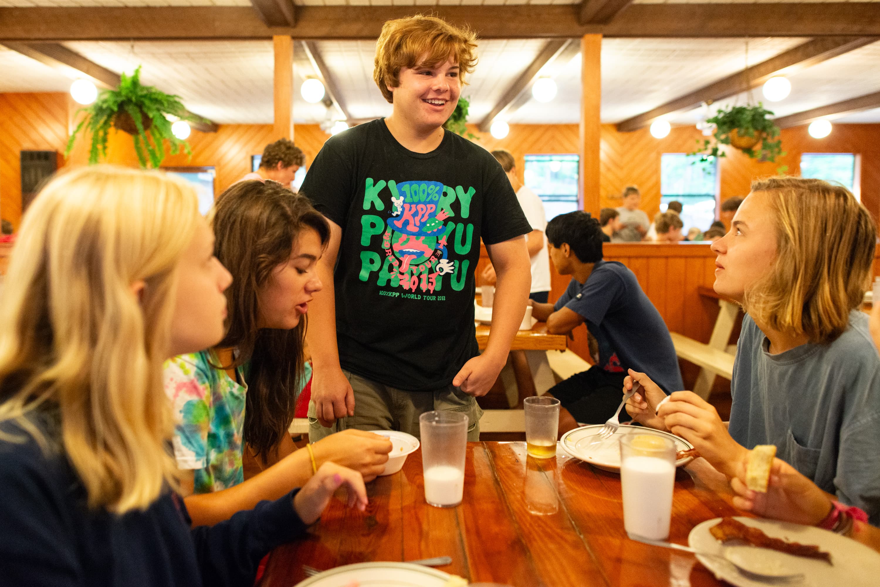 A group of young people sitting at a wooden table in a dining hall. One person is standing, wearing a T-shirt with colorful lettering. Others are seated with plates of food and drinks in front of them. Hanging plants and people at tables are visible in the background.