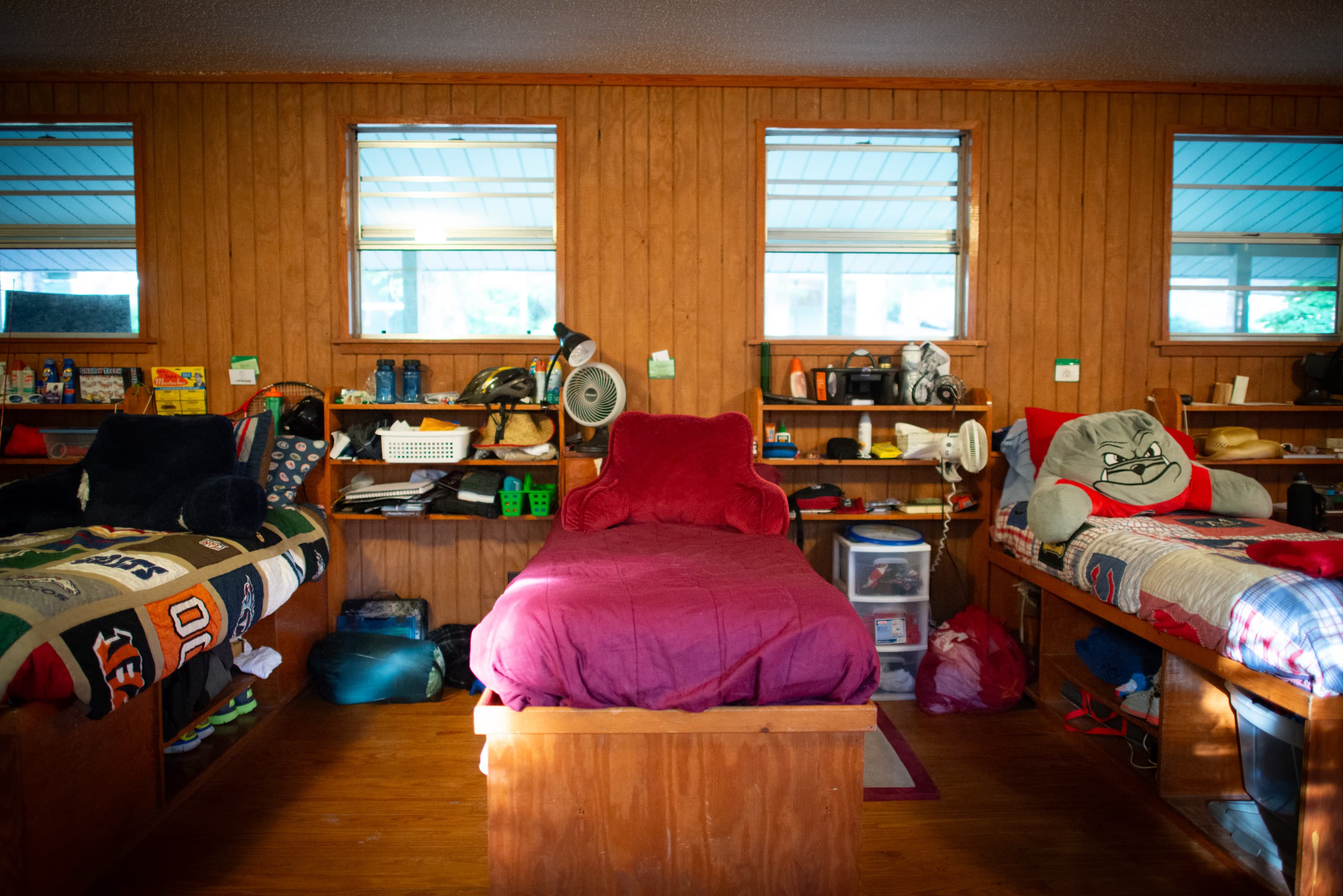 A room with three beds, each with its own bedding and personal items. The room has wood-paneled walls and three windows. Shelves above the beds hold various items like helmets, water bottles, and fans. The center bed has a burgundy cover and a backrest pillow. The bed on the right features a pillow with a cartoon face, and the bedding on the left has sports-themed patterns.