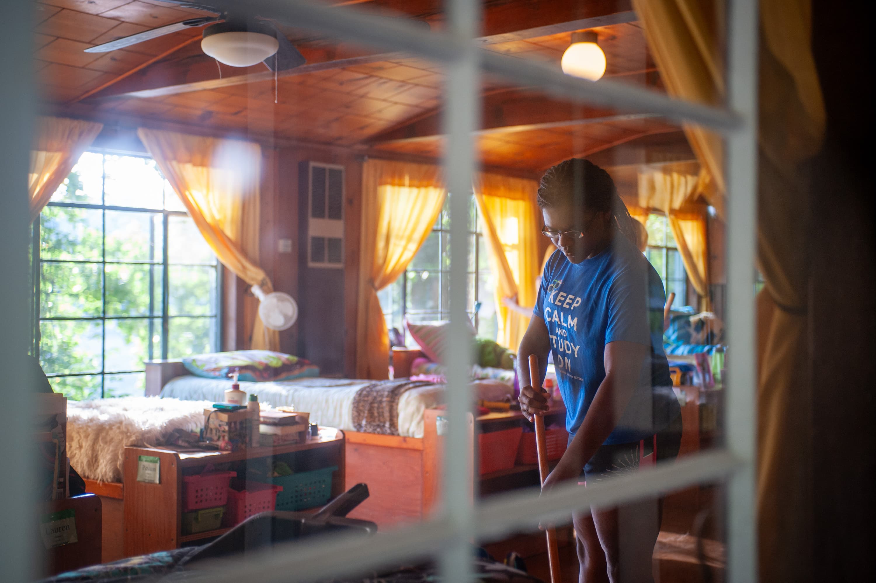 View through a window of a person inside a wooden room, sweeping the floor. The room has large windows with yellow curtains and beds with various items on shelves underneath.