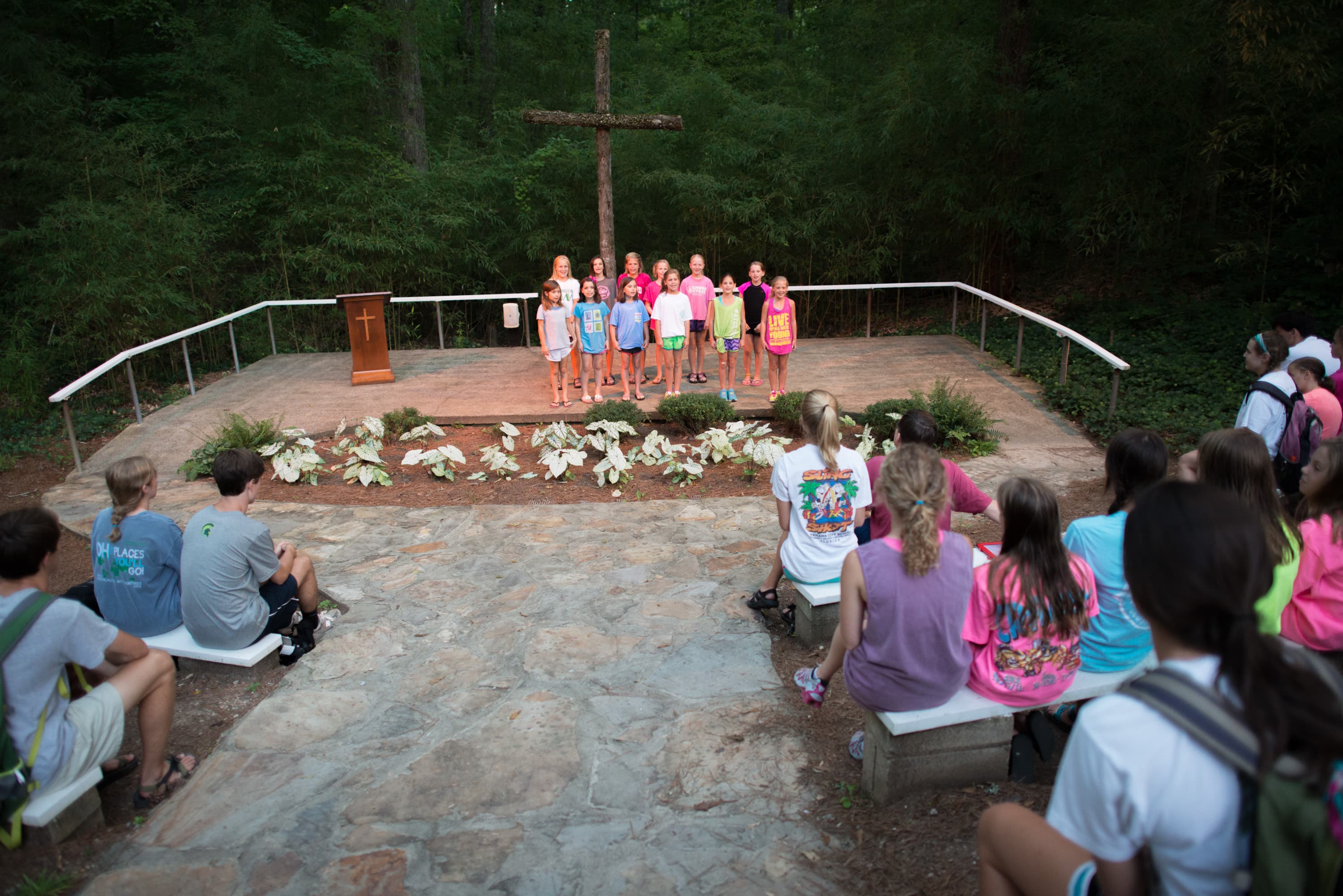 A group of children standing on a stone stage with a wooden cross in the background, surrounded by trees. An audience of seated children watches them. A wooden podium with a cross on it is on the stage.